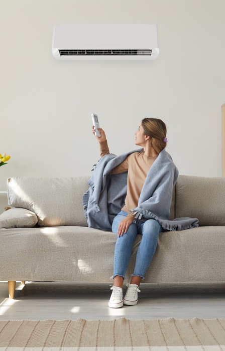 A female homeowner sitting on her living room sofa. She's turned around with a remote adjusting her AC unit on the wall.