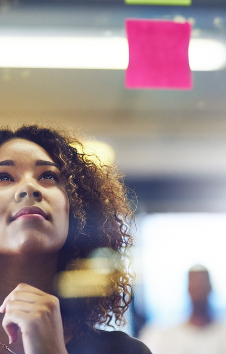 A woman looks thoughtfully at colorful sticky notes on a glass wall as she carefully plans creative ideas.