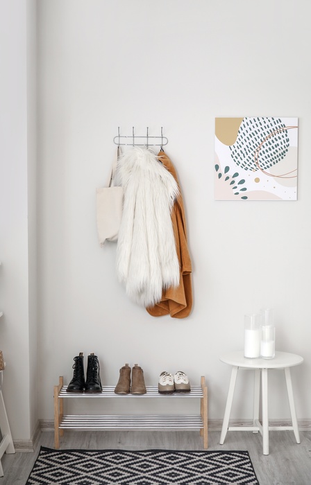 The entryway of a home with white walls, hooks holding jackets and purses, a shoe rack, and a white stool.
