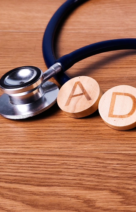 A hand placing a round wooden D tile beside ADHD letter tiles and a stethoscope on a wooden tabletop.