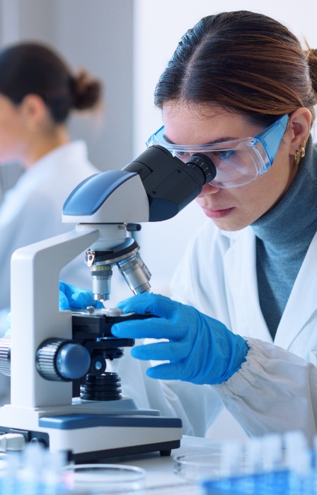 A woman wearing safety goggles and blue gloves using a microscope in a bright laboratory with another person nearby.