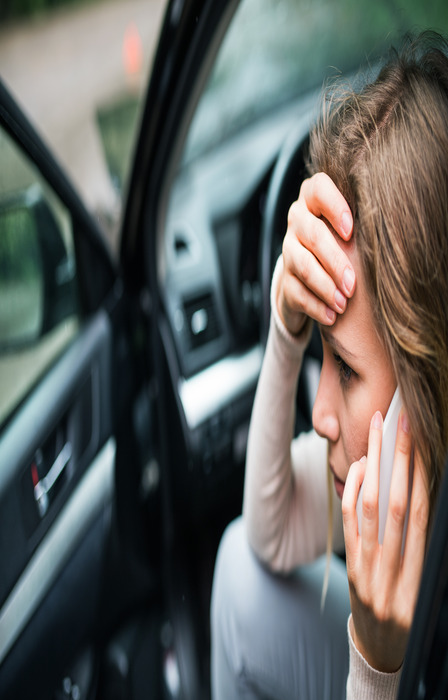 A young woman sits inside an open car with her hand on her forehead and a cell phone pressed to her ear.