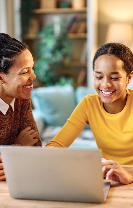 Woman and girl smile while looking at a laptop together at a home table, with a smartphone and pencil case nearby.