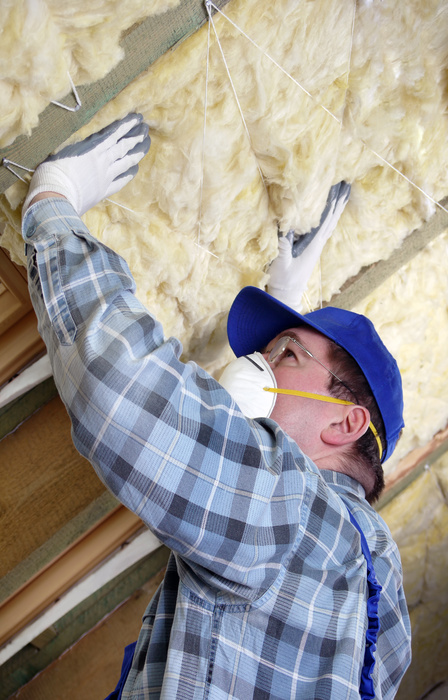 A person wearing gloves, a plaid long-sleeved shirt, and a mask is placing yellow insulation on a wall in an attic.
