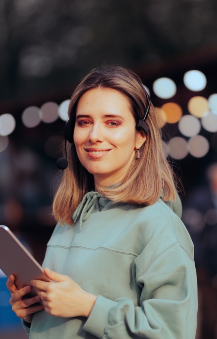 A woman with a production headset and a tablet standing in front of colorful bokeh lights at an outdoor event.