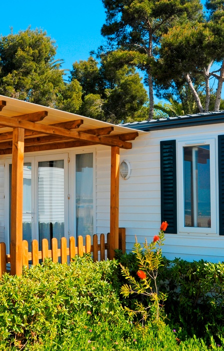 A modular home in a mobile community is surrounded by neatly trimmed bushes and features a wooden canopy.