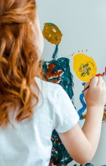 A red-headed girl holding a brush and paint palette is painting colorful flowers using an artist's easel.