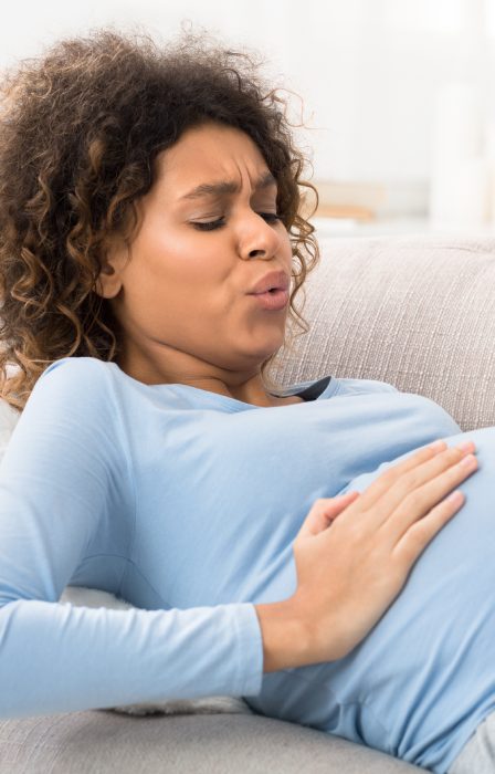 A woman wearing a blue long-sleeve shirt is lying on a couch with a look of pain on her face. She is holding her belly.