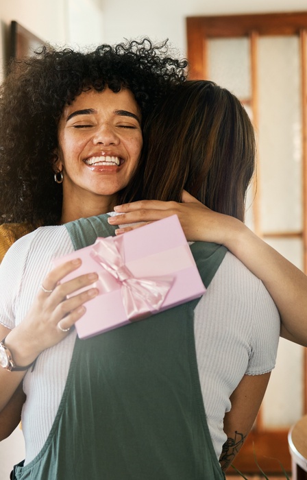 A woman smiles excitedly as she hugs another woman at home, while holding a gift wrapped with a pink ribbon.
