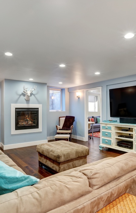 A modern finished basement features wooden flooring, a brown sectional, a mounted television, and a painted dresser.