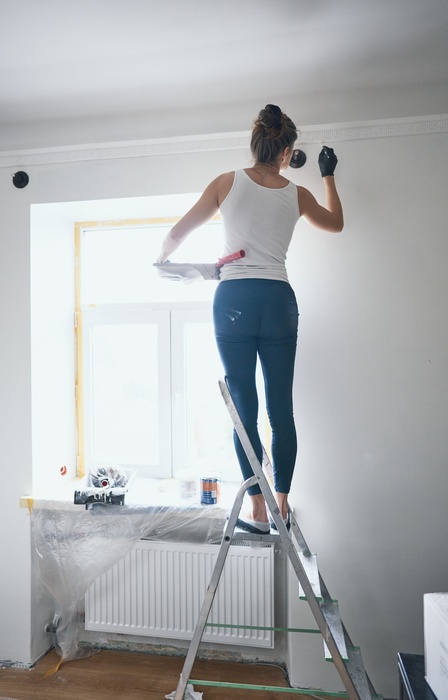 A woman stands on a ladder and uses a small brush to paint a wall in her home. She holds a paint tray with her other arm.