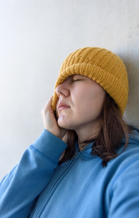 An anxious woman in a blue pullover tugs a yellow beanie over her right eye and leans against a white wall.