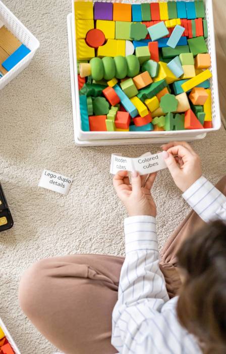 A woman sits on a white area rug surrounded by bins of toys. A label maker sits in front of her, and she holds several labels.