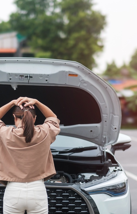 The back of a woman looking under the hood of her car. She has her hands on her head in frustration.