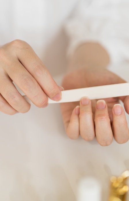 A close-up view shows a woman wearing a white shirt filing the nail on their ring finger with a white nail file.