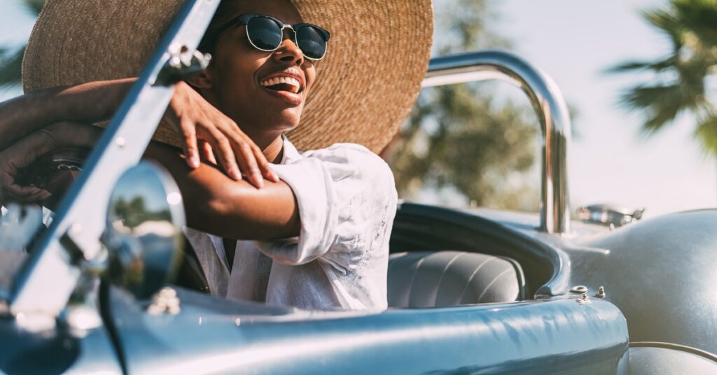 Four smiling women walking arm-in-arm outdoors. Three wear glasses, and two are in wide-brimmed straw hats.