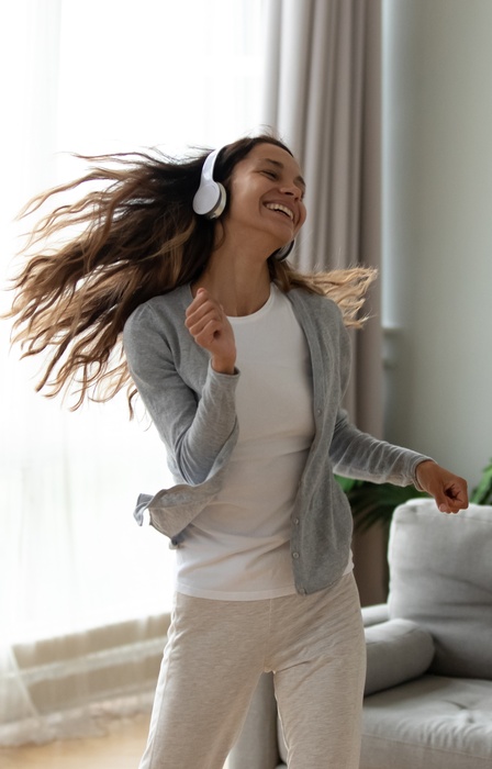 A young woman with long brunette hair wears a pair of white headphones as she dances while smiling widely.