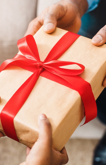 One pair of hands giving a wrapped present to another pair of hands. The gift is wrapped in brown paper and red ribbon.