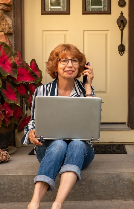 A woman sits on her front porch next to a large plant. She holds a laptop in her lap and has a phone next to her ear.
