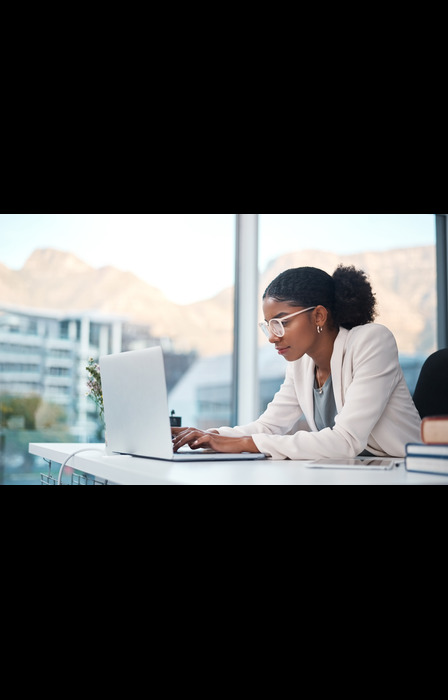 A focused young woman wearing glasses and a blazer typing on a laptop while working at a desk near a large picture window.