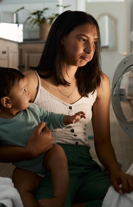 A young woman sits on the floor in front of a washing machine while holding a toddler and looking frustrated.