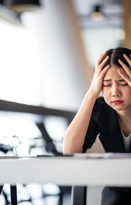A woman sits at her desk and looks stressed out. Her eyes are closed and she has her hands on her head.
