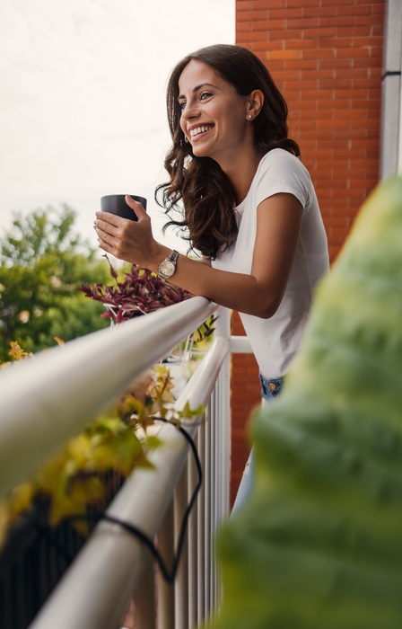 A woman stands on her balcony holding a coffee mug and smiles as she looks out over trees and greenery in the distance.