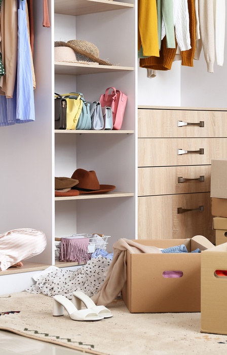 A young woman sitting in a large closet wearing a hat, with boxes of clothing stacked up and clothing scattered around.