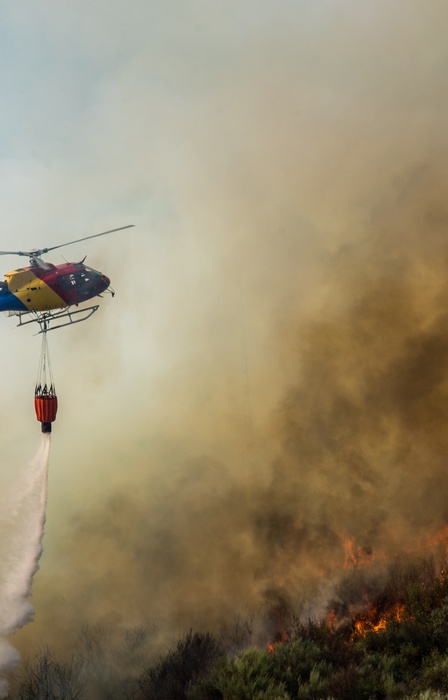 A rescue helicopter flying over a wildfire zone with a water tank dangling from the chopper. There are flames and smoke.