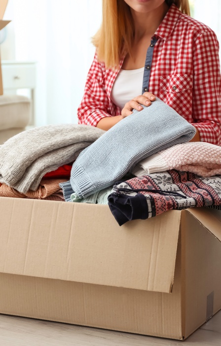 A woman sorts folded clothing into a cardboard donation box while another labeled donation box sits nearby.