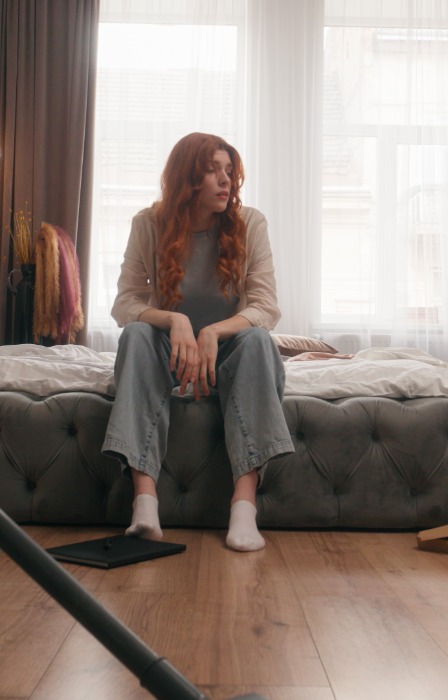 A woman sits on the edge of an unmade bed in a cluttered bedroom, surrounded by household items and a vacuum cleaner.
