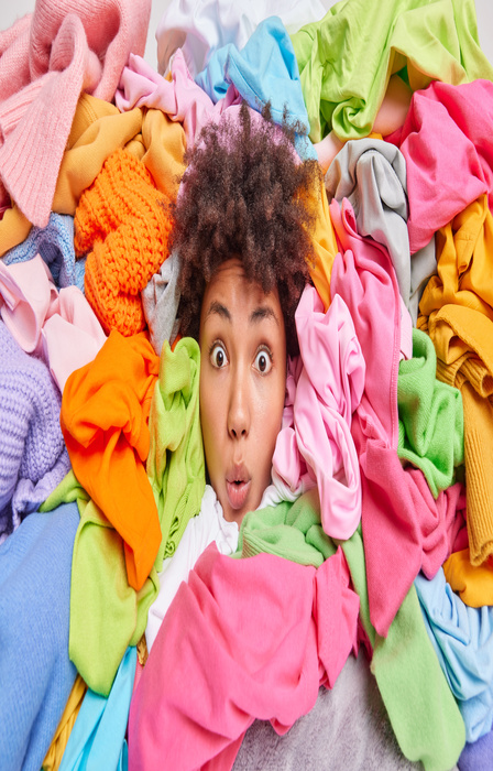 A surprised woman’s face peeks out from a vibrant pile of colorful clothes in pink, green, yellow, and blue hues.