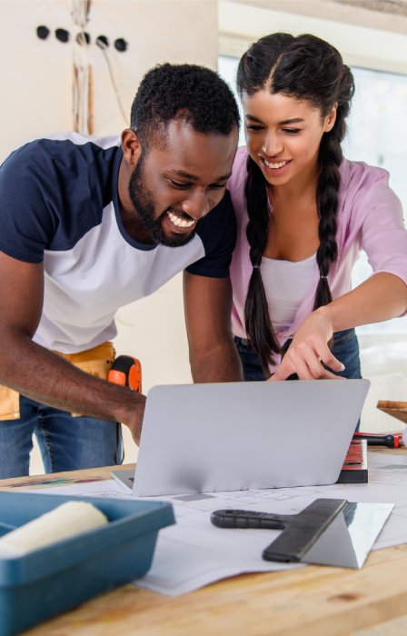 A man and woman look over a laptop set on a full work table. They grin, excited to work on their home expansion.