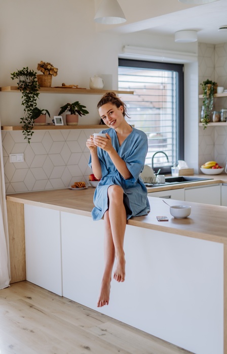 A woman in a robe sitting on a kitchen counter holding a mug, smiling in a bright kitchen with plants and shelves.