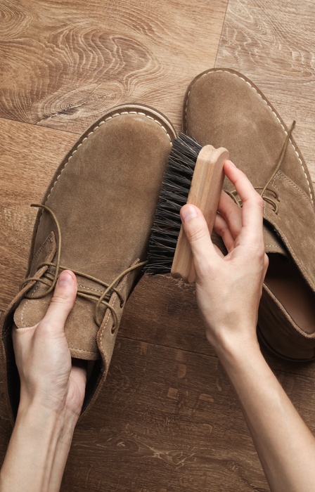 A person brushes a pair of brown leather shoes on a wooden floor as part of a simple at-home care routine.