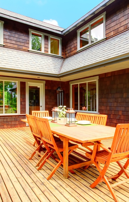 A wooden deck connected to the corner of a house. There is a large dining space in the middle, but the rest is quite open.