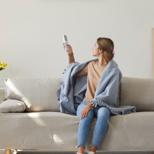 A female homeowner sitting on her living room sofa. She's turned around with a remote adjusting her AC unit on the wall.