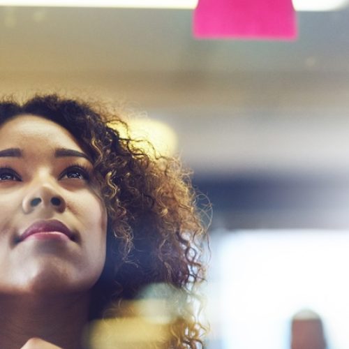 A woman looks thoughtfully at colorful sticky notes on a glass wall as she carefully plans creative ideas.