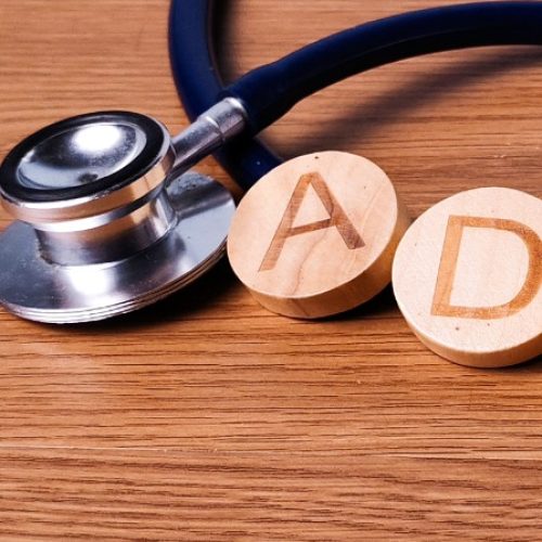 A hand placing a round wooden D tile beside ADHD letter tiles and a stethoscope on a wooden tabletop.