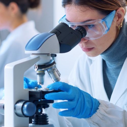 A woman wearing safety goggles and blue gloves using a microscope in a bright laboratory with another person nearby.