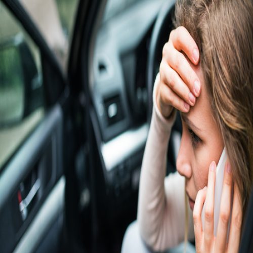 A young woman sits inside an open car with her hand on her forehead and a cell phone pressed to her ear.