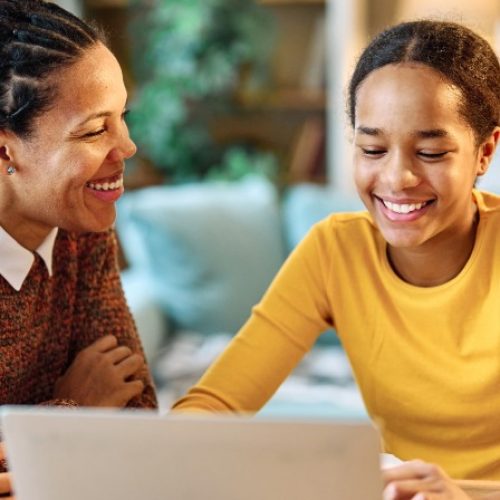 Woman and girl smile while looking at a laptop together at a home table, with a smartphone and pencil case nearby.