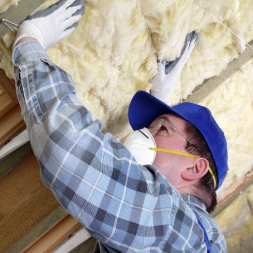A person wearing gloves, a plaid long-sleeved shirt, and a mask is placing yellow insulation on a wall in an attic.