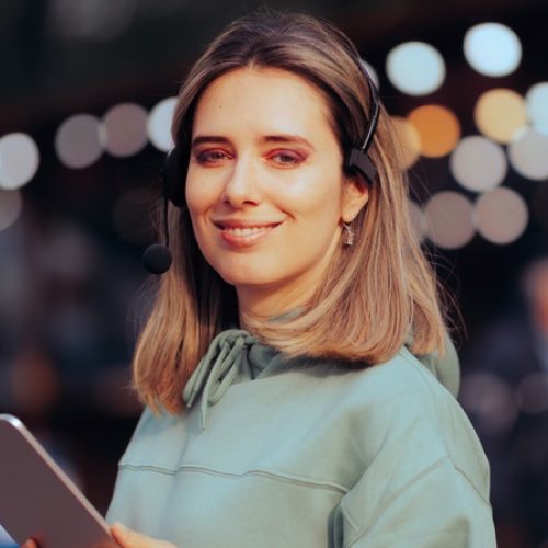A woman with a production headset and a tablet standing in front of colorful bokeh lights at an outdoor event.