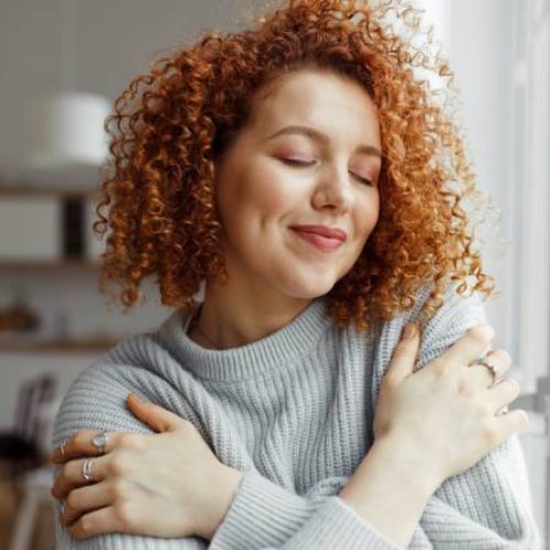 A woman with red curly hair smiles and hugs herself next to a window, enjoying the texture of her cozy pullover.