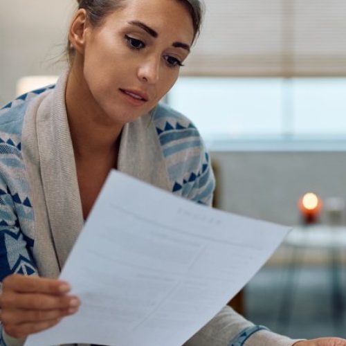 A woman sitting next to a counter holds up a sheet of paper and reads it. She holds more papers in her other hand.
