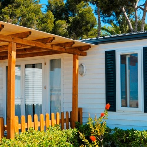 A modular home in a mobile community is surrounded by neatly trimmed bushes and features a wooden canopy.