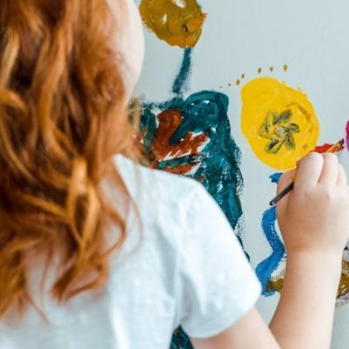 A red-headed girl holding a brush and paint palette is painting colorful flowers using an artist's easel.