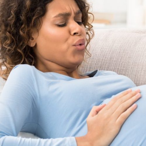 A woman wearing a blue long-sleeve shirt is lying on a couch with a look of pain on her face. She is holding her belly.
