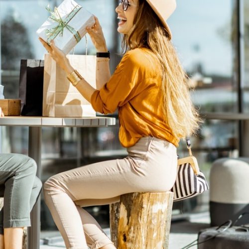 Two smiling women wearing hats sit at an outdoor café table, and one is happily giving the other a gift.